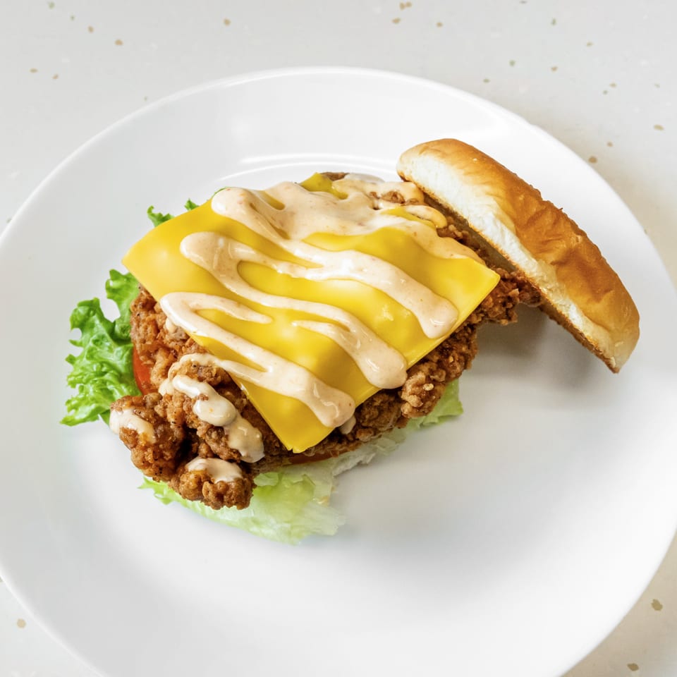 Fried Shack's fried chicken sandwich with melted cheese, sauce, lettuce, and tomato on a white plate.