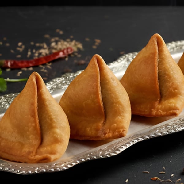 Four golden-brown samosas arranged on a silver platter with spices and a red chili in the background.